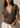A woman wearing a dark brown short-sleeve V-neck t-shirt and off-white jeans, standing against a textured beige wall with warm sunlight and shadows.