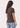 A woman with long wavy brown hair is shown from the back wearing a dark brown short-sleeve crewneck t-shirt and light blue jeans against a plain white background.